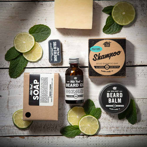 The Mod Cabin soap, beard oil, beard balm and solid cologne laid out on a white rustic wood table surrounded by slices of bergamot and mint leaves. All are packaged in glass, aluminum or paper packaging.